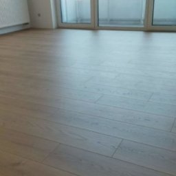 Paweł Mroszczyk - Jasne panele podłogowe ułożone w pustym pokoju z białymi ścianami i grzejnikiem. Widok na balkon przez przeszklone drzwi.