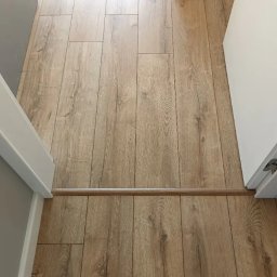 Paweł Mroszczyk - Ułożone panele podłogowe w odcieniach drewna, widok z góry, z listwą progową między pomieszczeniami. Jasne ściany i futryny drzwi.