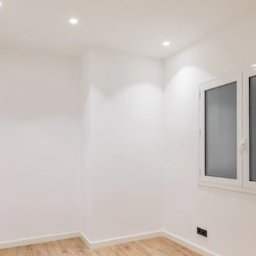 Paweł Mroszczyk - Jasne wnętrze po remoncie: białe ściany, drewnopodobna podłoga, okno z matową szybą i minimalistyczne oświetlenie punktowe. Widok na odnowiony pokój.