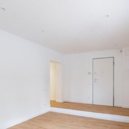 Paweł Mroszczyk - Jasne, puste wnętrze po remoncie: świeżo pomalowane ściany, panele podłogowe, białe drzwi i okno z roletą. Minimalistyczny design, czystość i porządek.