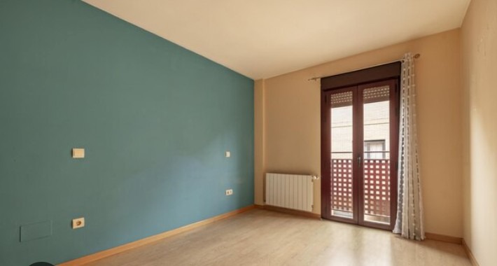 Pusty pokój po remoncie: turkusowa ściana z gniazdkami, jasne panele podłogowe, grzejnik pod oknem balkonowym z brązowymi okiennicami i zasłoną.