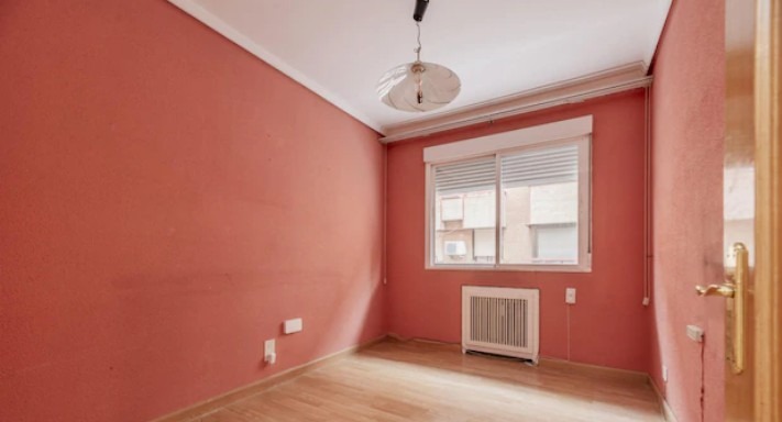 Pokój z pomarańczowymi ścianami i drewnianą podłogą. Widoczne okno z żaluzjami, grzejnik i lampa sufitowa. Ściany mają wyraźną fakturę.