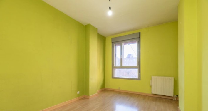 Pusty pokój z zielonymi ścianami, drewnianą podłogą i grzejnikiem pod oknem z roletą. Widoczna żarówka na suficie. Świeżo pomalowane ściany w odcieniu limonki.