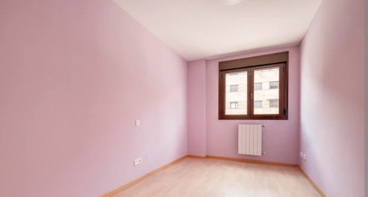 Pusty pokój po malowaniu ścian na delikatny, pudrowy róż. Jasna podłoga, okno z brązową ramą i biały grzejnik. Widoczne gniazdka elektryczne.
