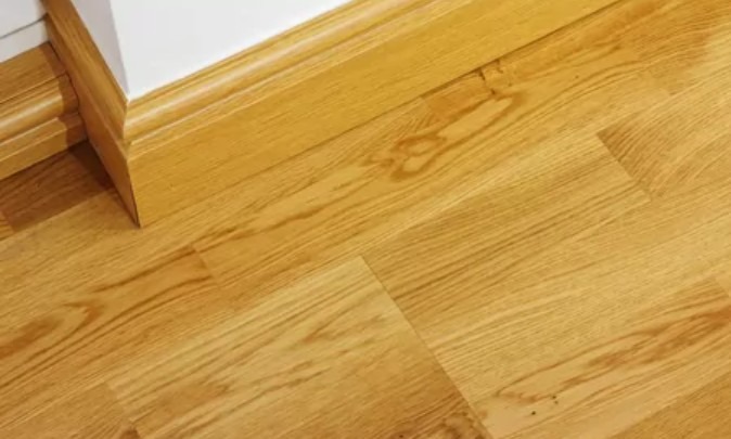 Jasne panele podłogowe ułożone w jodełkę, z widoczną przypodłogową listwą. Naturalne odcienie drewna i subtelne usłojenie. Kątowe ujęcie podkreśla wzór.