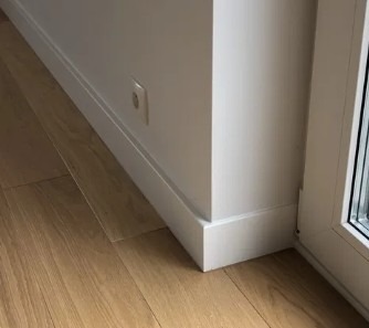 Biała listwa przypodłogowa na styku ściany i jasnej, drewnianej podłogi. Widoczny fragment gniazdka elektrycznego i ramy okiennej. Minimalistyczne wykończenie wnętrza.