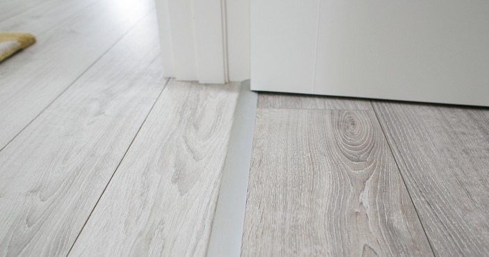 Jasnoszare panele podłogowe z widoczną listwą progową i fragmentem białych drzwi. Delikatny wzór drewna, gładka powierzchnia, minimalistyczny detal wykończeniowy.