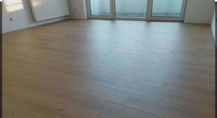 Jasne panele podłogowe ułożone w pustym pokoju z białymi ścianami i grzejnikiem. Widok na balkon przez przeszklone drzwi.