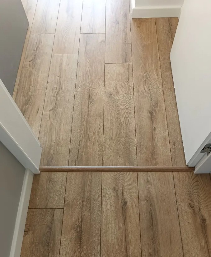 Ułożone panele podłogowe w odcieniach drewna, widok z góry, z listwą progową między pomieszczeniami. Jasne ściany i futryny drzwi.