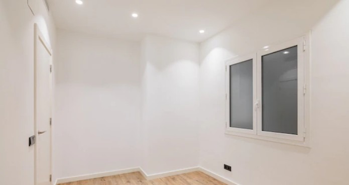 Jasne wnętrze po remoncie: białe ściany, drewnopodobna podłoga, okno z matową szybą i minimalistyczne oświetlenie punktowe. Widok na odnowiony pokój.