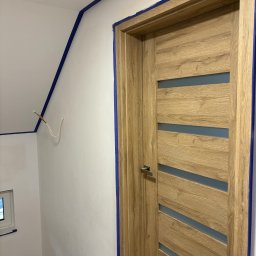Miłosz Bartelak - Świeżo pomalowane białe ściany z niebieską taśmą malarską wzdłuż krawędzi, drewniane drzwi z poziomymi przeszkleniami, wystające przewody elektryczne.