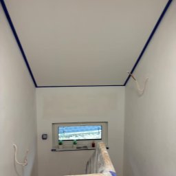 Miłosz Bartelak - Przygotowane do malowania wnętrze: świeżo zagruntowane ściany i sufit zabezpieczone niebieską taśmą malarską, widoczne wystające kable i osłonięte folią schody.