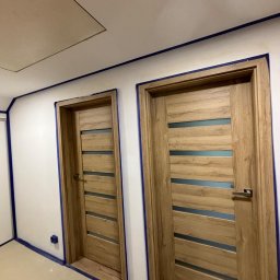 Miłosz Bartelak - Dwa drewniane drzwi wewnętrzne z poziomymi przeszkleniami, zabezpieczone taśmą malarską przed malowaniem ścian, widoczne ślady po pracach remontowych na podłodze i suficie.