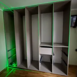 Montaż szafy wnękowej z użyciem lasera krzyżowego, widoczne poziome i pionowe linie lasera, drewniana podłoga.