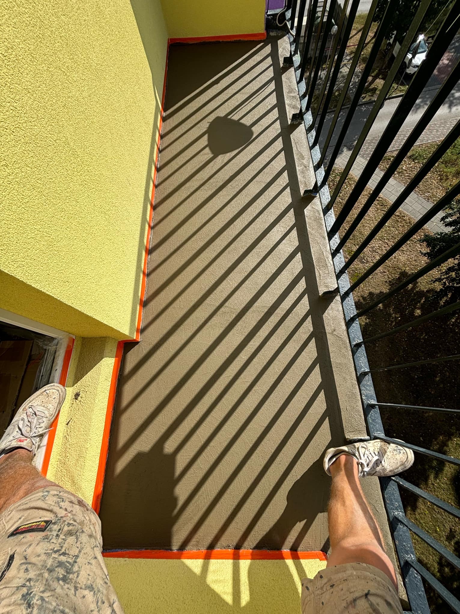 Świeżo wylana, wyrównana wylewka na balkonie, zabezpieczona pomarańczową taśmą, z widocznymi cieniami barierki i fragmentami obuwia osoby robiącej zdjęcie.