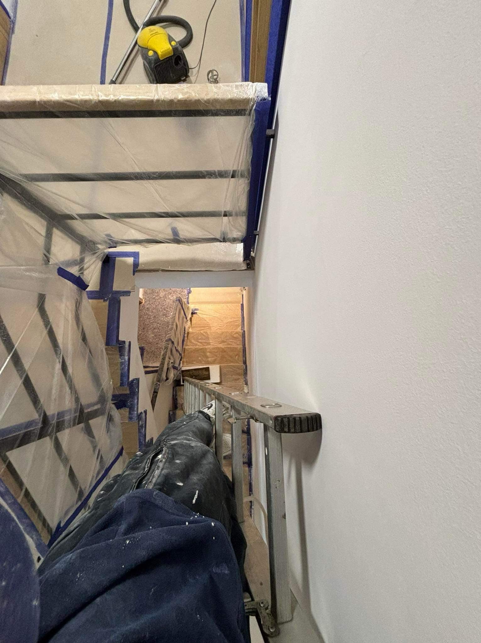 Świeżo pomalowana biała ściana, widok z góry z drabiny, zabezpieczone folią i taśmą malarską elementy, widoczny odkurzacz przemysłowy na górze rusztowania.