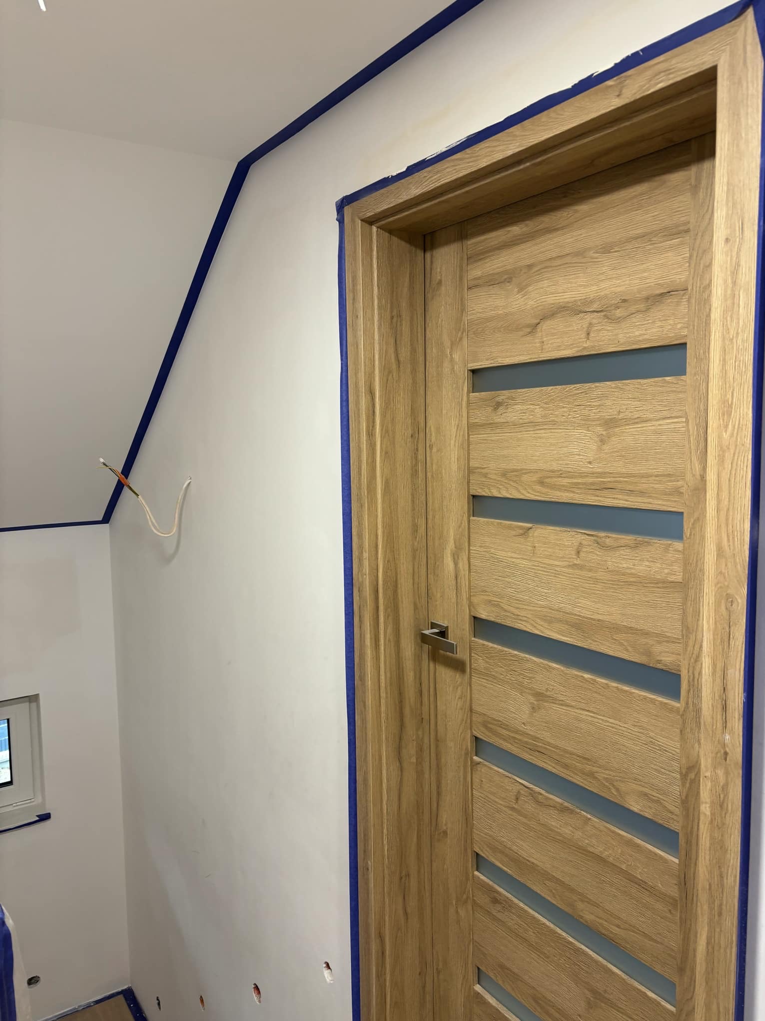 Świeżo pomalowane na biało ściany z niebieską taśmą malarską, drewniane drzwi z przeszkleniami, wystające kable elektryczne.