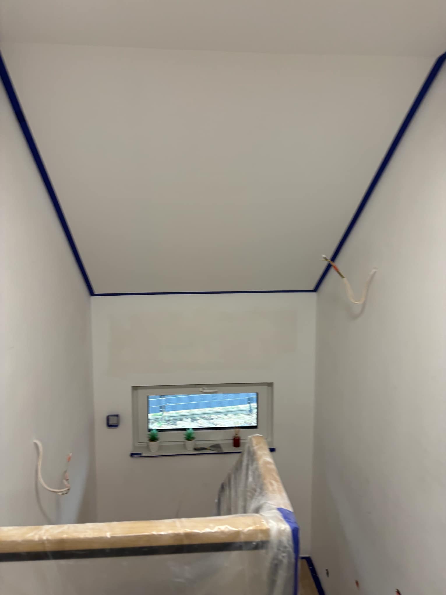 Przygotowane do malowania wnętrze: świeżo zagruntowane ściany i sufit zabezpieczone niebieską taśmą malarską, widoczne wystające kable i osłonięte folią schody.