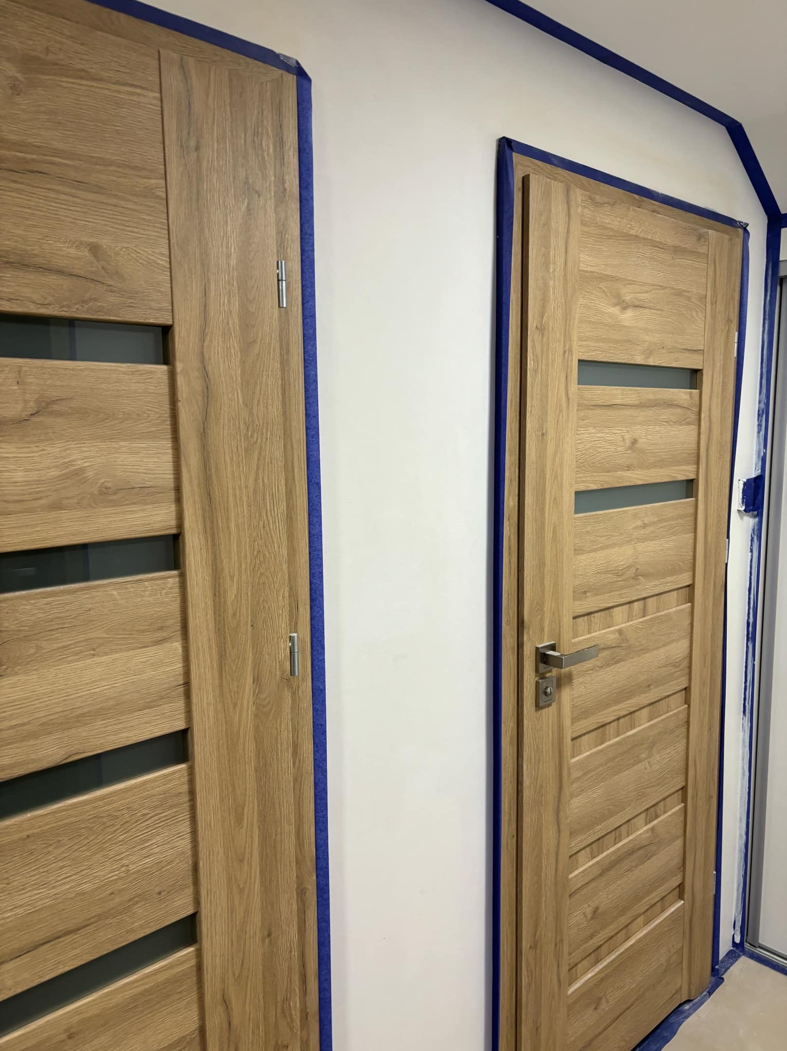 Dwa drewniane drzwi wewnętrzne z przeszkleniami, zabezpieczone taśmą malarską przed malowaniem ścian.