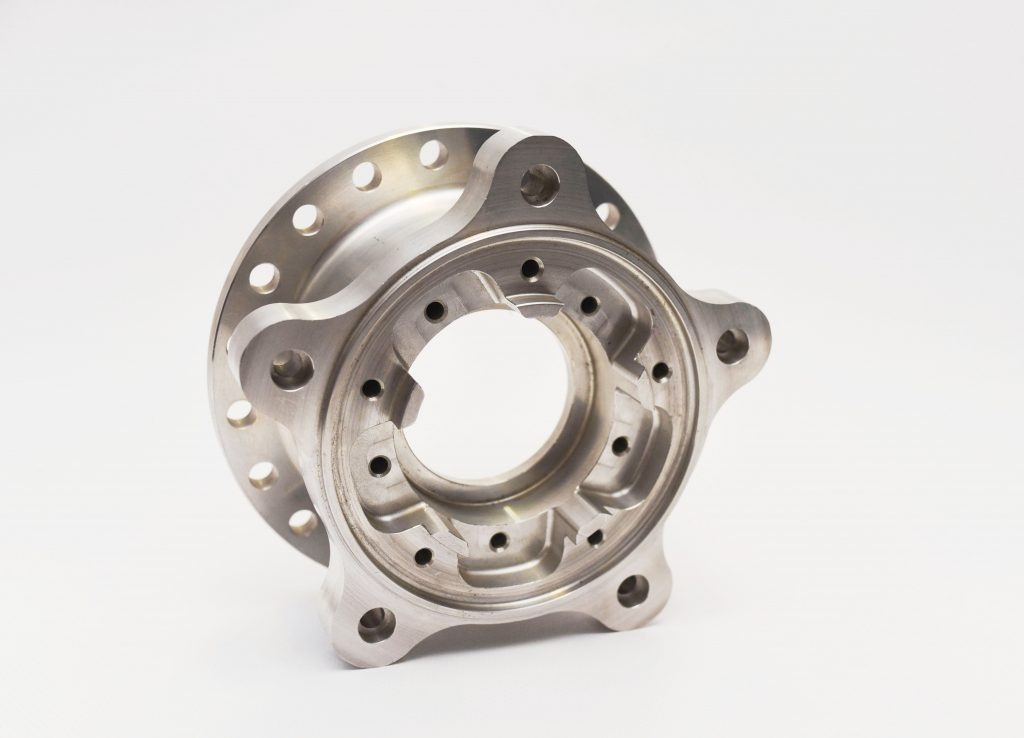 Precyzyjnie wykonany, metalowy element z licznymi otworami i zagłębieniami, obrobiony na jasny, matowy kolor, ustawiony pod kątem na białym tle, ukazujący skomplikowaną geometrię wewnętrzną.