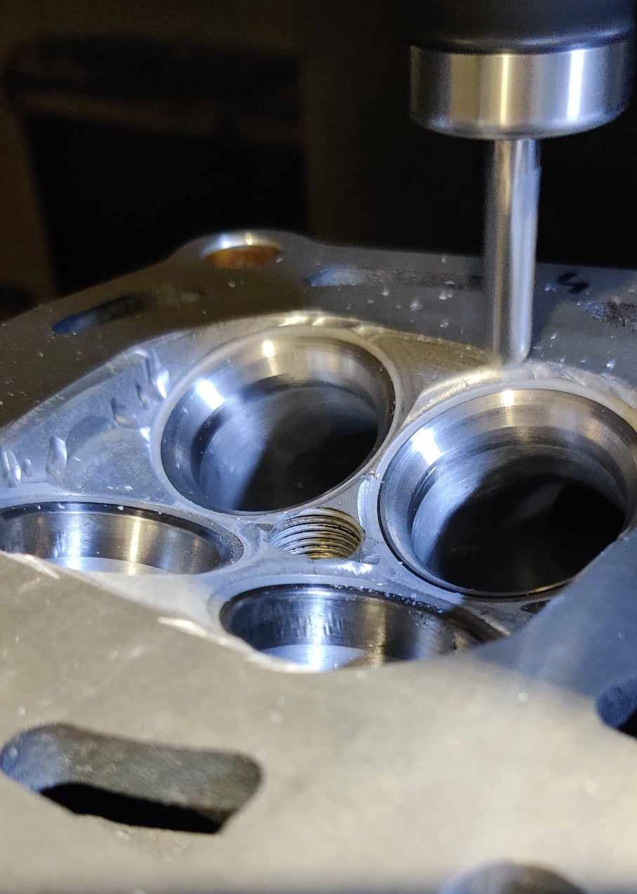 Frezarka CNC obrabia aluminiową głowicę silnika spalinowego, widoczne otwory zaworowe i gwintowany otwór.