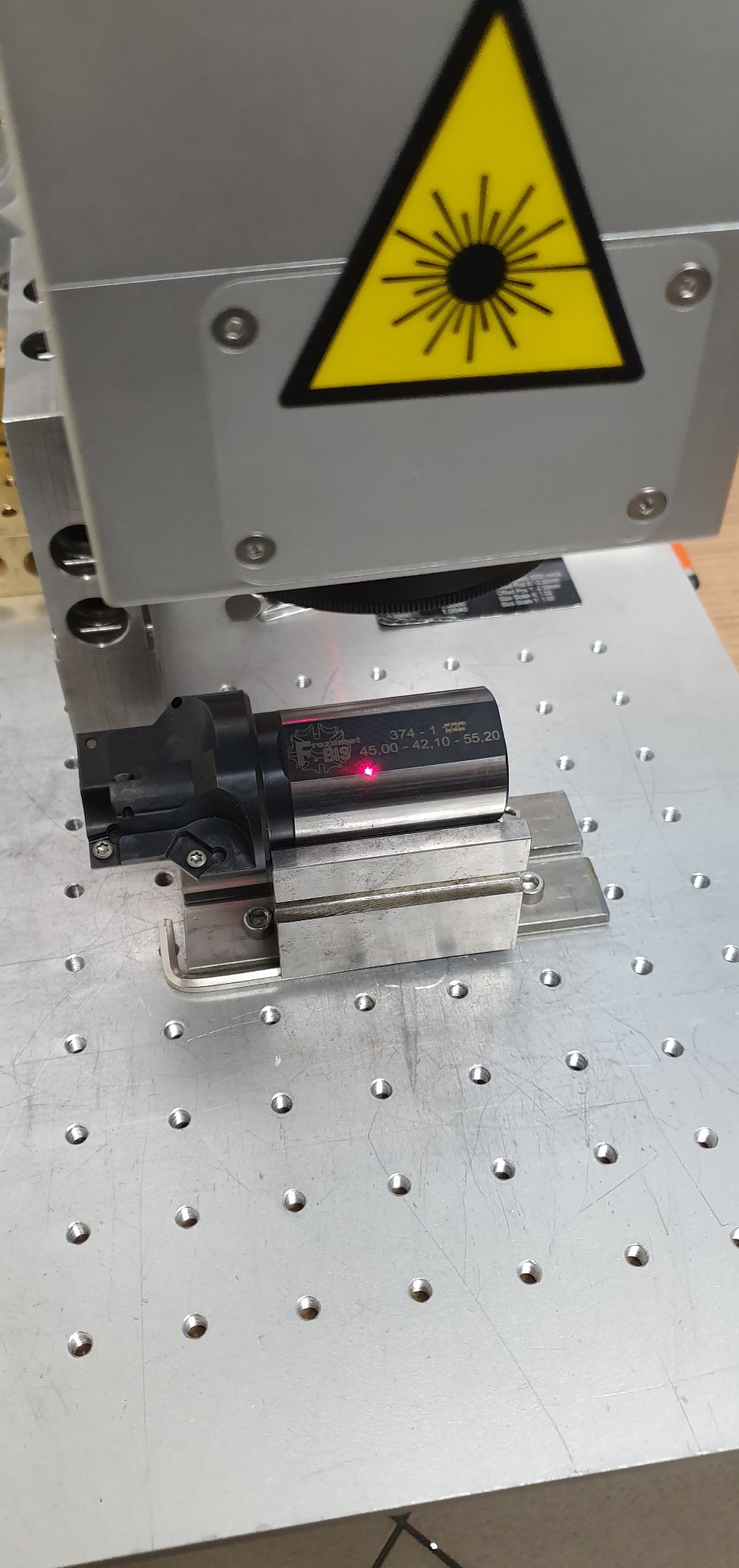 Proces znakowania laserowego elementu z tworzywa sztucznego w maszynie CNC, widoczny czerwony punkt lasera i żółty znak ostrzegawczy.