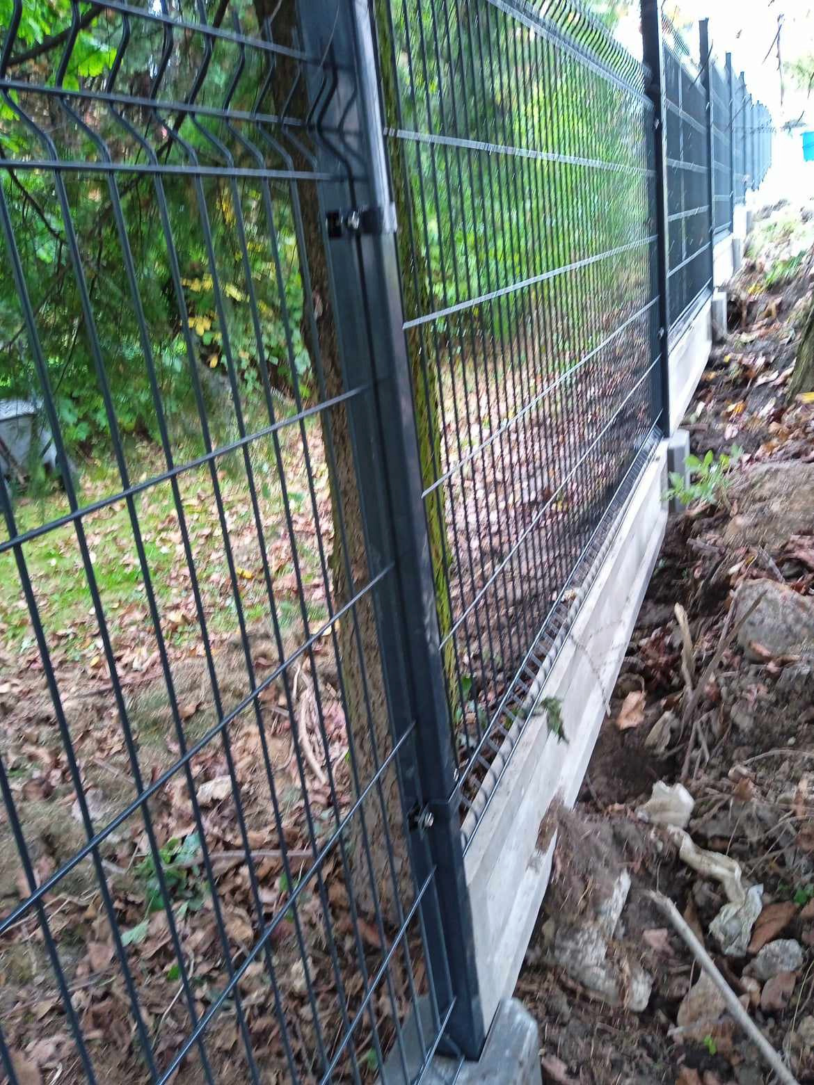 Nowoczesne ogrodzenie panelowe z podmurówką betonową, widok z boku, z drzewem w tle.