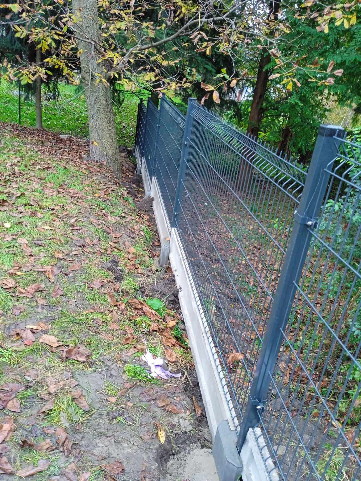 Nowoczesne, antracytowe ogrodzenie z paneli na betonowej podmurówce, zamontowane na nierównym terenie z drzewami i opadłymi liśćmi, widoczne elementy montażowe.