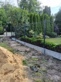 Montaż ogrodzenia z siatki: słupki osadzone w betonowej podmurówce, widoczny fragment siatki i pryzma piasku na pierwszym planie.