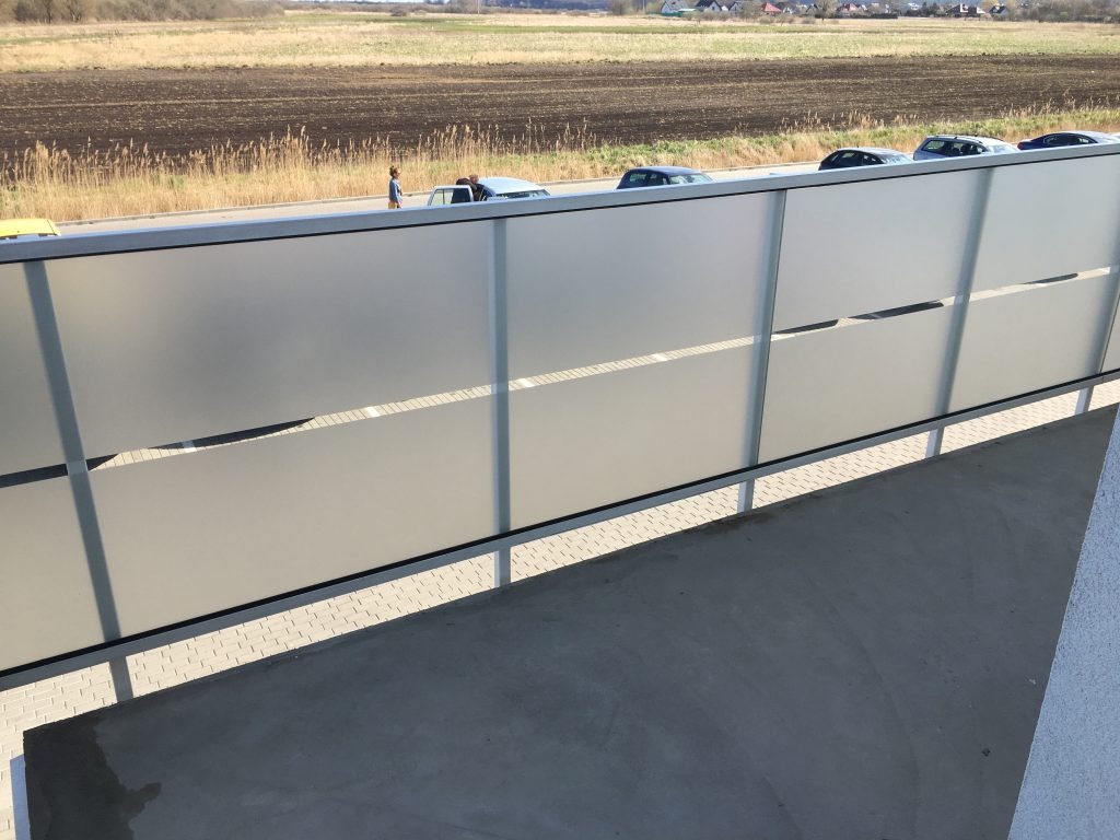 Dźwiękochłonny ekran akustyczny wzdłuż drogi z widocznymi samochodami i osobą. Tło stanowią pola i zabudowania.