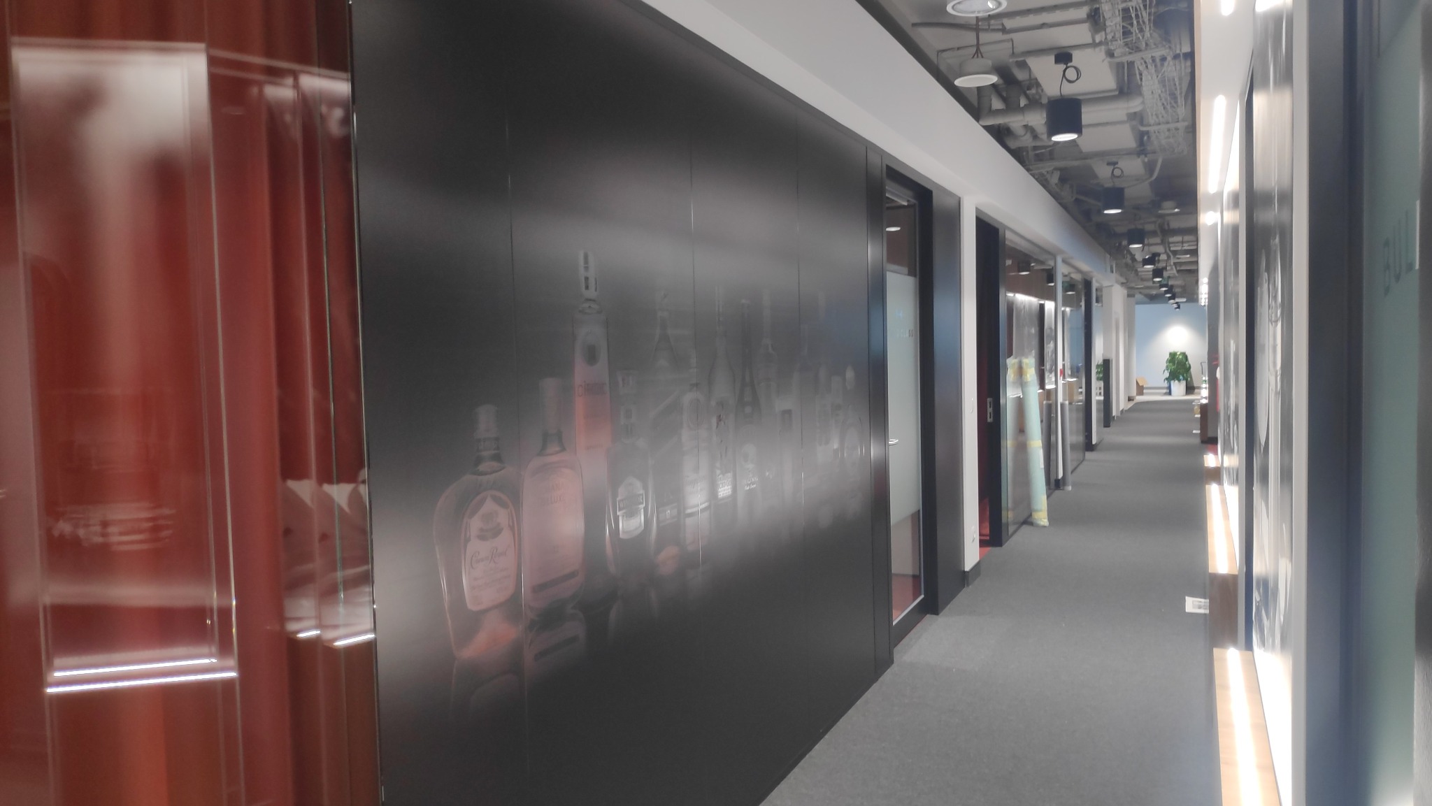 Długi korytarz biurowy z czarną ścianą ozdobioną grafiką przedstawiającą butelki różnych alkoholi w odcieniach szarości, widoczne oświetlenie sufitowe i szklane ściany po obu stronach.