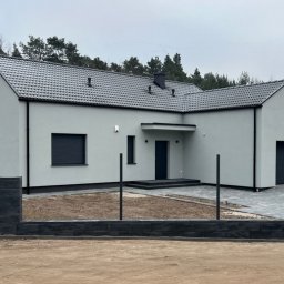 PRZEDSIĘBIORSTWO BUDOWLANE IWANOW SPÓŁKA Z OGRANICZONĄ ODPOWIEDZIALNOŚCIĄ - Nowoczesny parterowy dom z szarą dachówką i elewacją, widoczny zza niskiego ogrodzenia z betonowych bloczków, z podjazdem wyłożonym kostką brukową, w tle las.