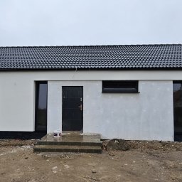PRZEDSIĘBIORSTWO BUDOWLANE IWANOW SPÓŁKA Z OGRANICZONĄ ODPOWIEDZIALNOŚCIĄ - Nowoczesny, parterowy budynek z czarną dachówką i oknami w ciemnych ramach, widoczny podczas prac wykończeniowych, z resztkami farby i wiadrem na schodach wejściowych.