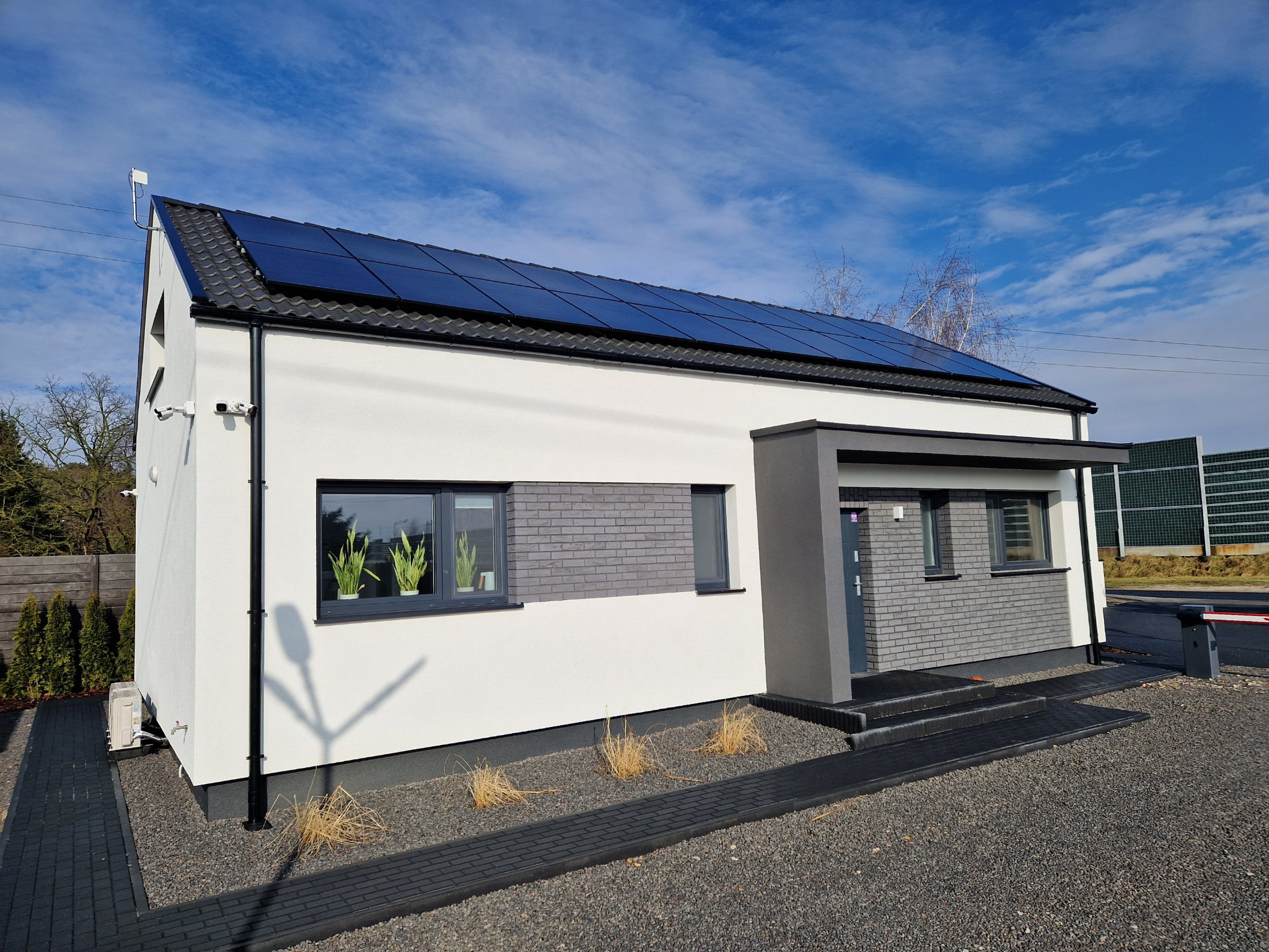 Nowoczesny dom jednorodzinny z panelami słonecznymi na dachu, szarym wykończeniem elewacji, oknami z roślinami i monitoringiem, otoczony szarym żwirem.