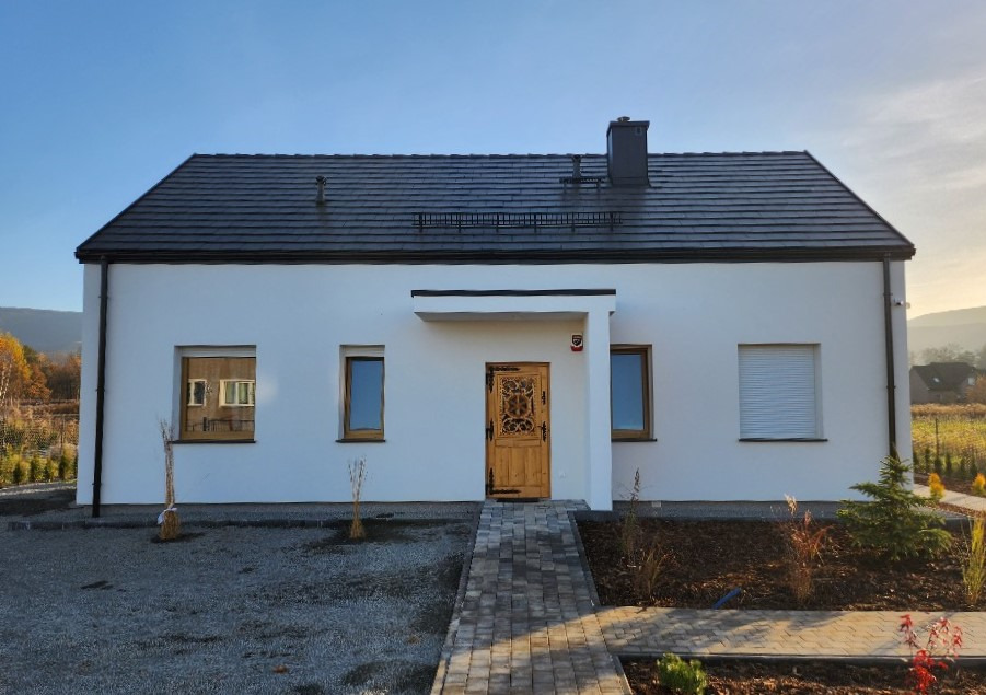 Nowoczesny dom z białymi ścianami, ciemnym dachem i ozdobnymi drewnianymi drzwiami wejściowymi, otoczony młodymi nasadzeniami i brukowanym chodnikiem.