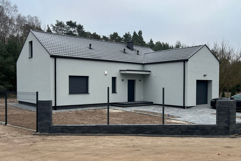 Nowoczesny parterowy dom z szarą dachówką i elewacją, widoczny zza niskiego ogrodzenia z betonowych bloczków, z podjazdem wyłożonym kostką brukową, w tle las.
