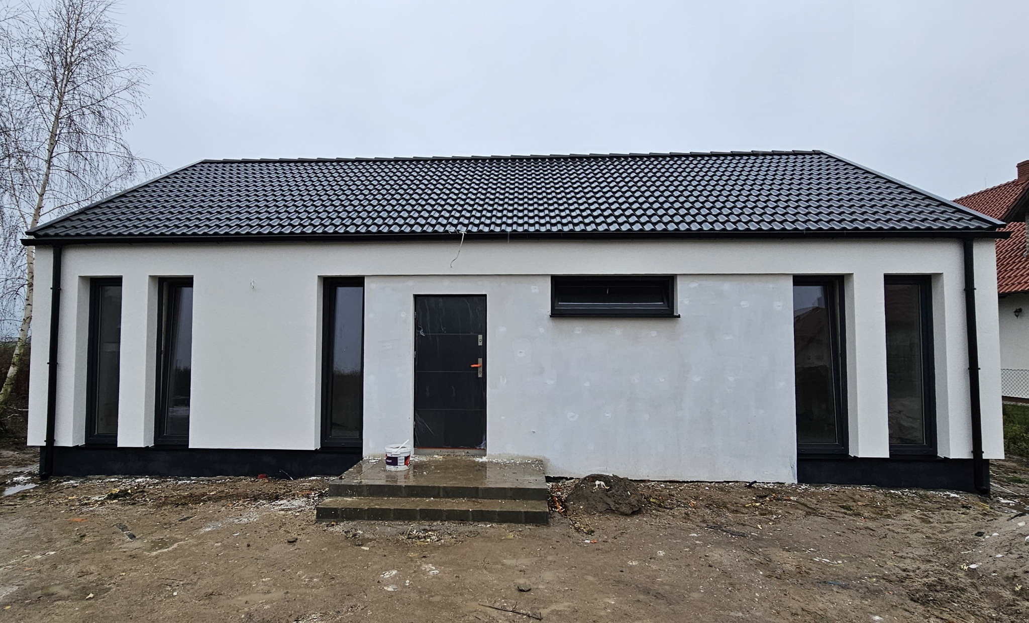 Nowoczesny, parterowy budynek z czarną dachówką i oknami w ciemnych ramach, widoczny podczas prac wykończeniowych, z resztkami farby i wiadrem na schodach wejściowych.