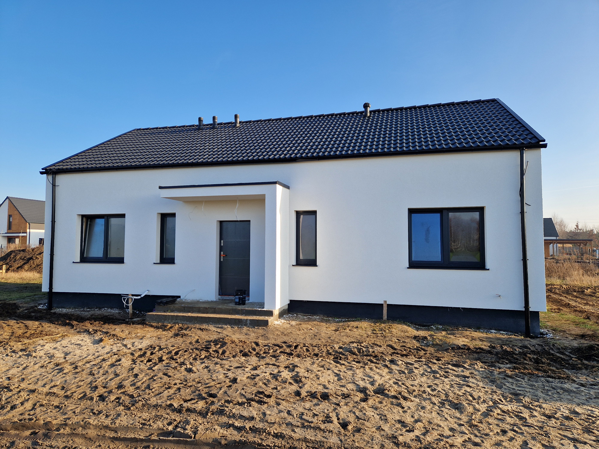 Nowoczesny, parterowy dom z białymi ścianami i czarnym dachem na tle błękitnego nieba, widoczne rury wentylacyjne na dachu oraz teren wokół domu w trakcie zagospodarowywania.