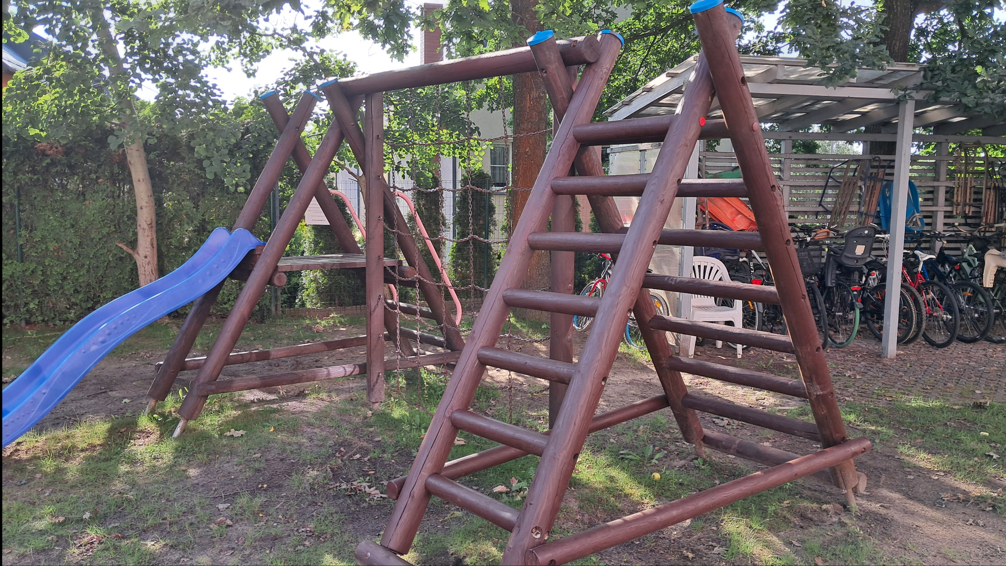 Drewniany plac zabaw z niebieską zjeżdżalnią, drabinką i siatką do wspinaczki, ustawiony na trawiastym terenie. W tle widoczne rowery pod zadaszeniem.