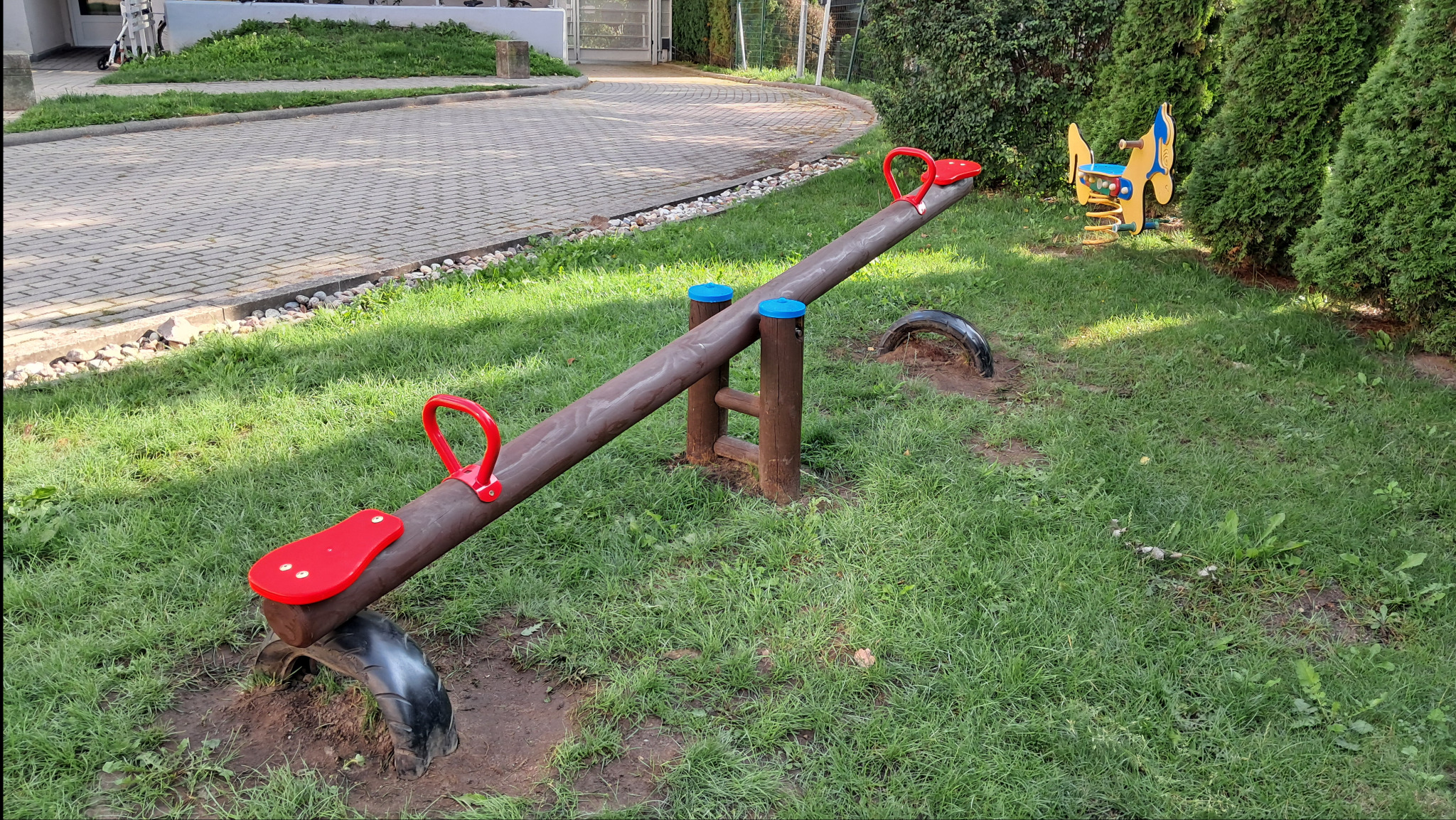 Drewniany plac zabaw z huśtawką na oponach i konikiem na sprężynie, otoczony zieloną trawą i tujami. Elementy w kolorach brązu, czerwieni i niebieskiego.