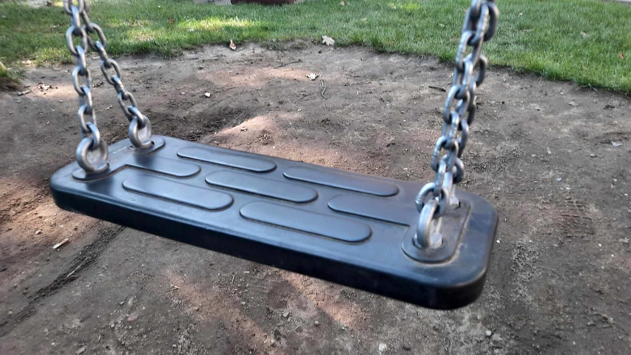 Czarne siedzisko huśtawki z łańcuchami na tle ziemi i trawy. Widok z dołu, ukazujący detale siedziska i mocowania łańcuchów.