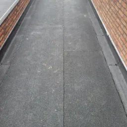 Płaski dach pokryty papą z widocznymi łączeniami, obrzeżony ceglanym murem i metalową listwą, z niewielkim kominkiem wentylacyjnym.