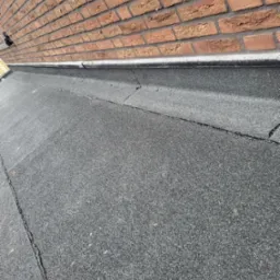 Płaski dach pokryty czarną papą bitumiczną z widocznymi pęknięciami i nierównościami, przylegający do ceglanej ściany budynku.