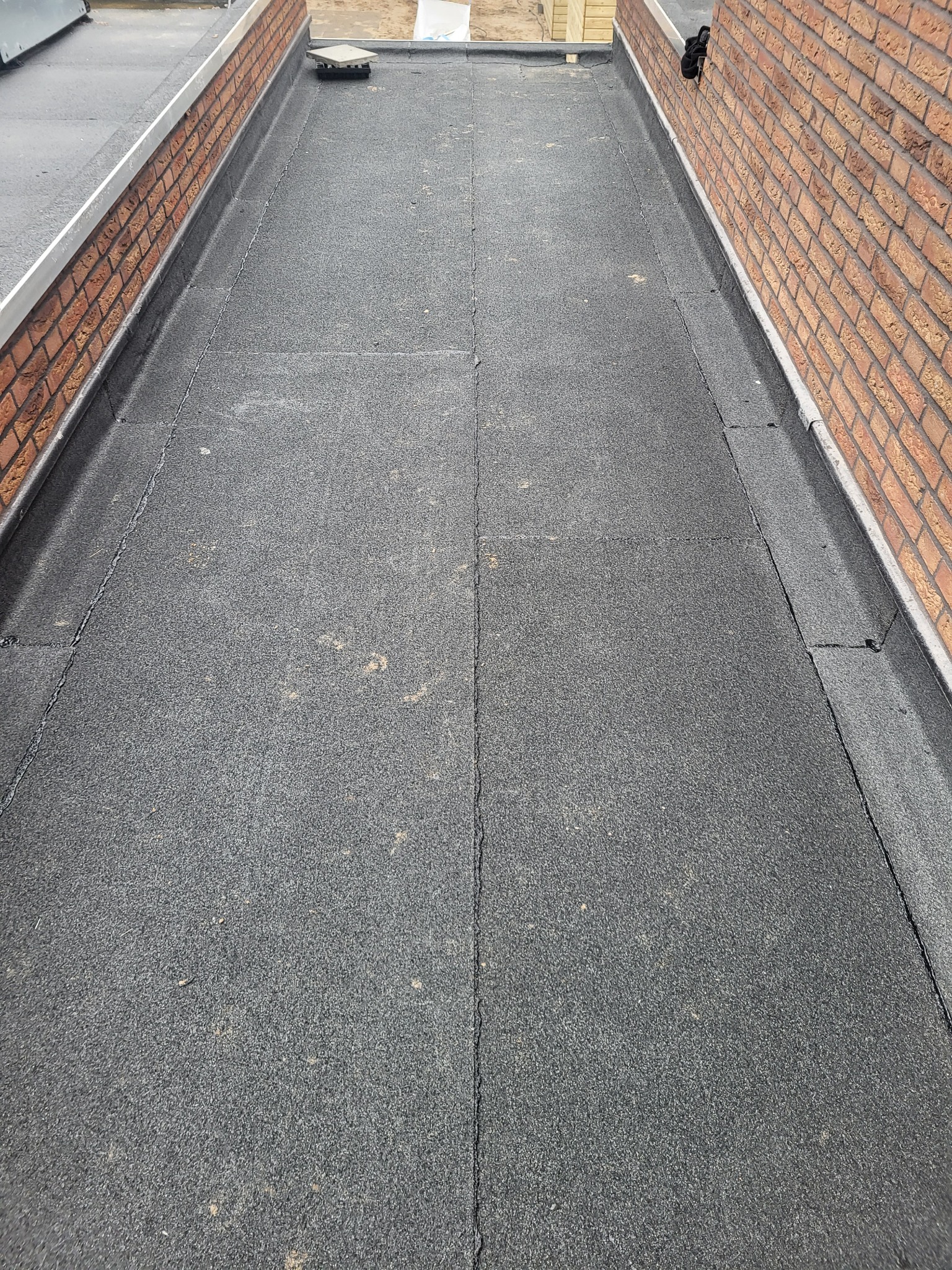 Płaski dach pokryty papą z widocznymi łączeniami, obrzeżony ceglanym murem i metalową listwą, z niewielkim kominkiem wentylacyjnym.