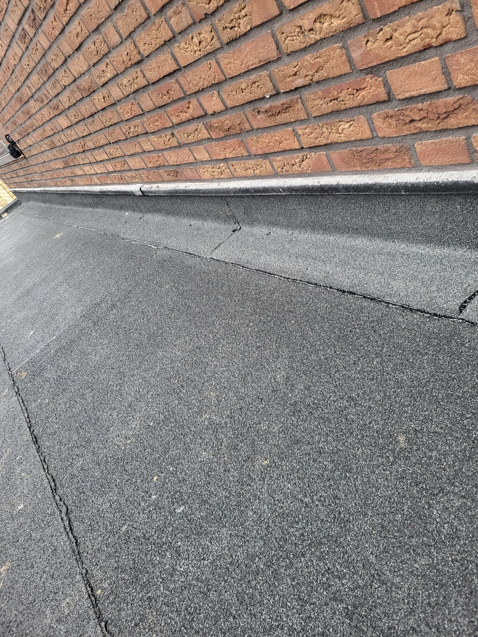 Płaski dach pokryty czarną papą bitumiczną z widocznymi pęknięciami i nierównościami, przylegający do ceglanej ściany budynku.