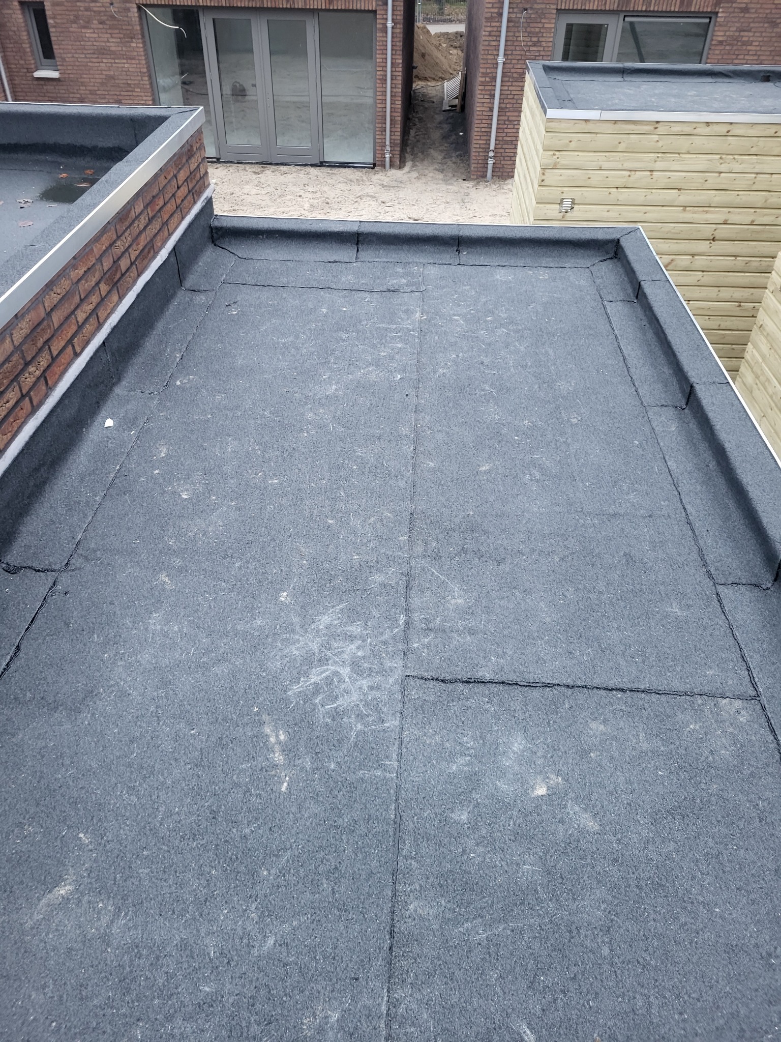 Wykończony płaski dach pokryty czarną papą, z obróbką blacharską na krawędziach i ceglaną ścianą attyki w tle.