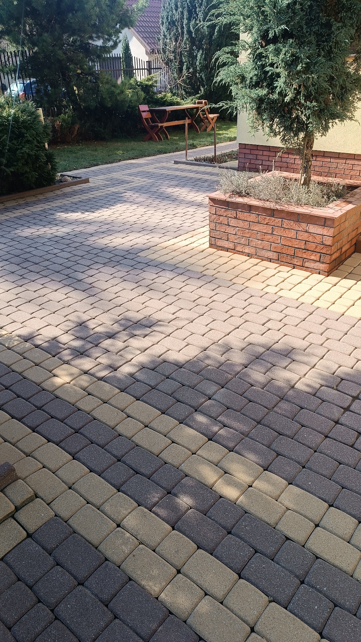 Ułożona kostka brukowa w odcieniach szarości i beżu tworząca wzór na podwórku, z widoczną donicą z cegły i zielenią w tle.