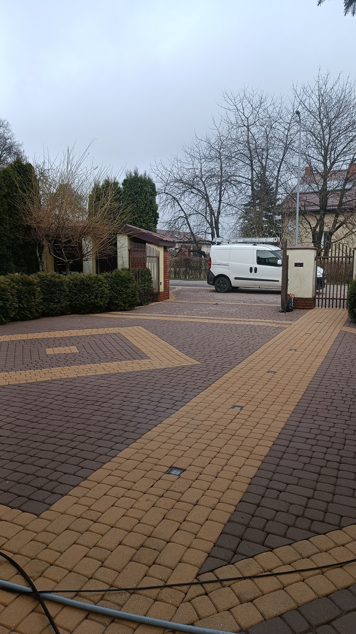 Dwukolorowa kostka brukowa ułożona we wzór na podjeździe, z białym vanem z drabinką na dachu w tle, otoczona zielenią i budynkami mieszkalnymi