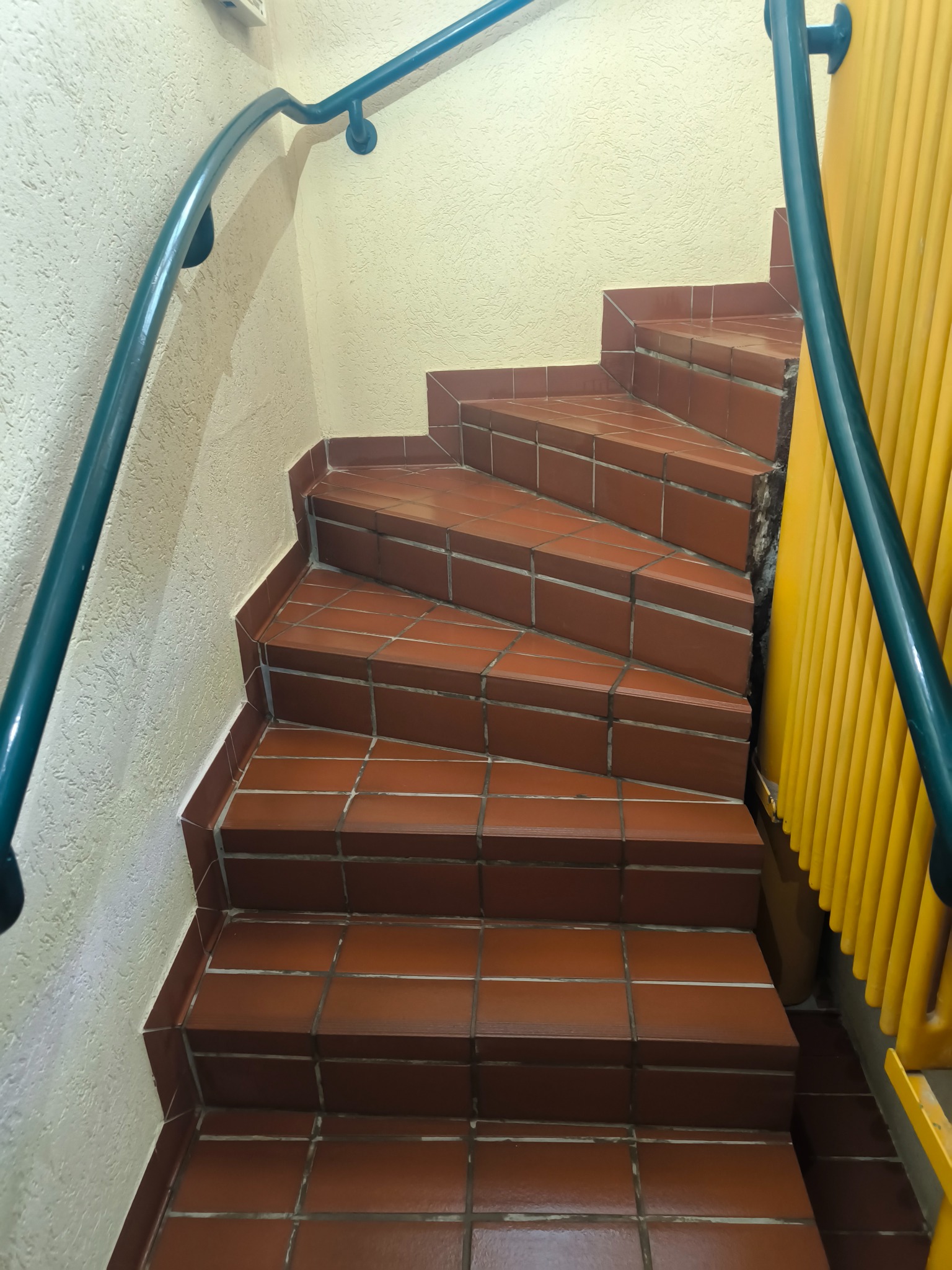 Klatka schodowa z brązowymi płytkami, niebieską poręczą i żółtym grzejnikiem. Widok z góry na kręte schody wewnątrz budynku. Teksturowana ściana w tle.