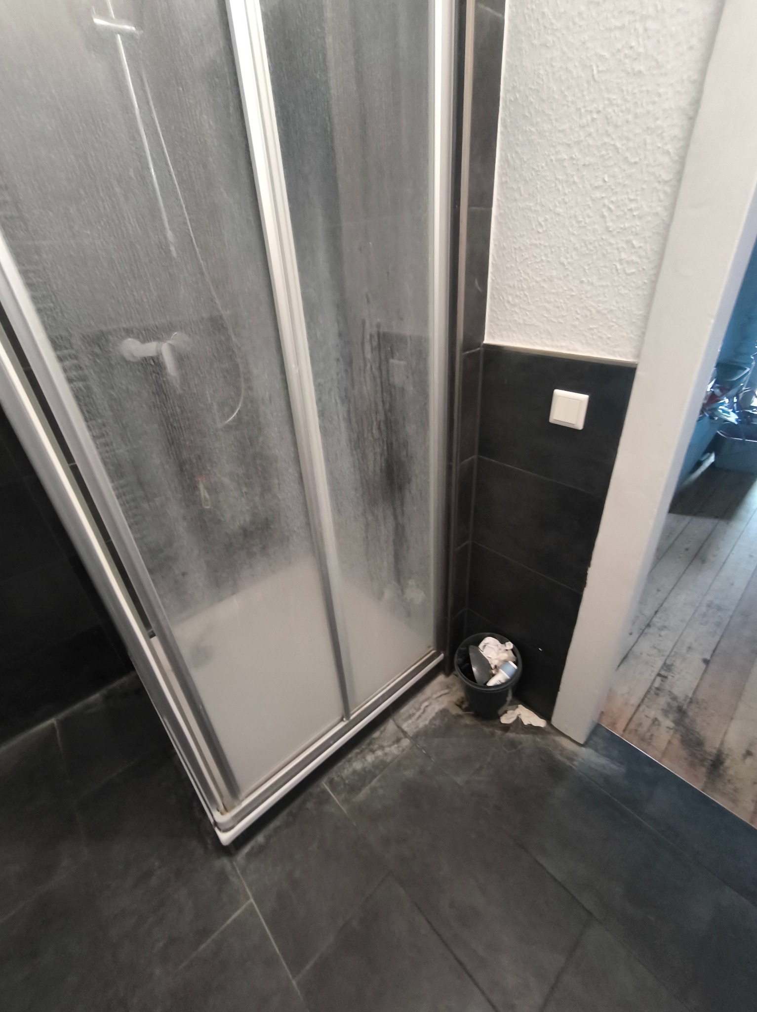 Zabrudzona kabina prysznicowa z widocznymi zaciekami i osadami, czarne płytki podłogowe i ściany, mały kosz na śmieci obok kabiny. Widok na fragment podłogi poza łazienką.
